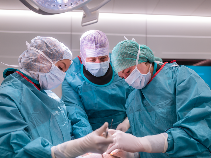 Prof. Thomas Dreher stands between Anna Schuler and a fellow resident and gives instructions on how to perform a paediatric procedure. All three people are wearing surgical clothing.