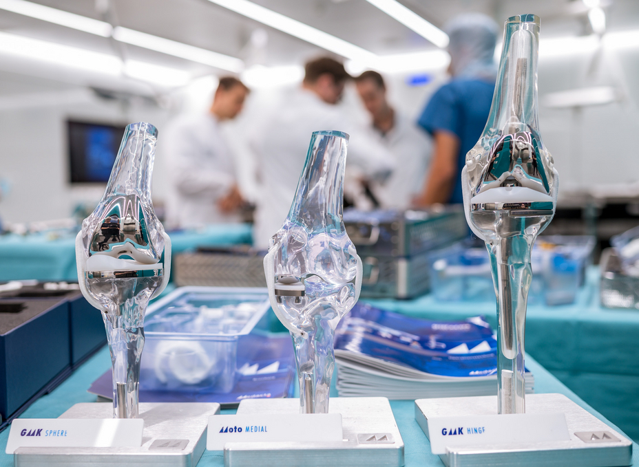 Three knee prosthesis models from Medacta stand on an instrument table in the OR-X, while a group of surgeons discuss in the background