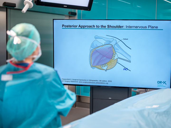Anna Schuler steht vor einem grossen TV-Screen auf dem eine Präsentationsslide mit Anweisungen zum Posterior Approach to the shoulder stehen.