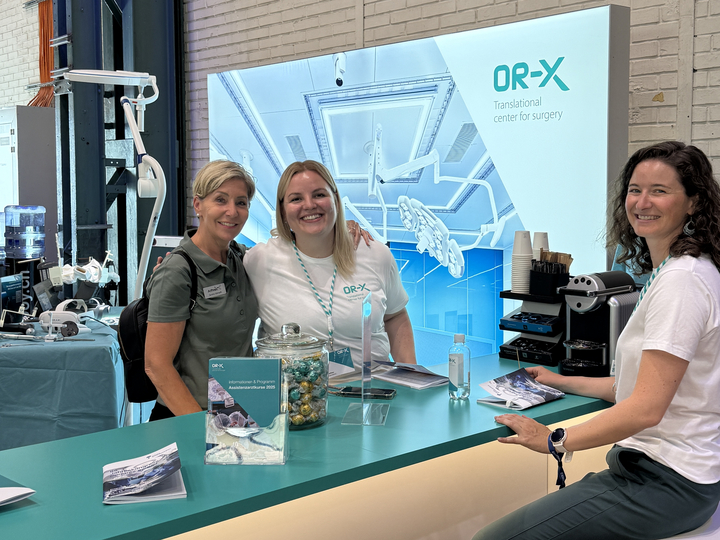 OR-X team members sit on high chairs with a visitor at the booth and smile into the camera.