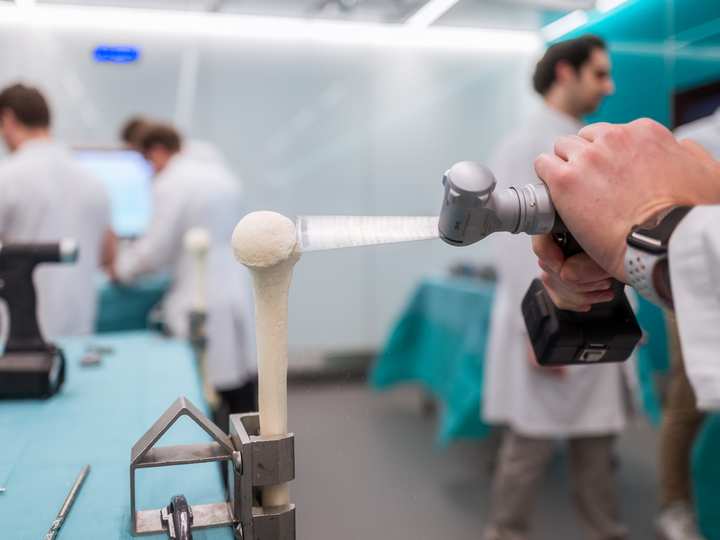 In the foreground is a close-up of an artificial femur being cut with a surgical saw. In the background are other residents working at their station.